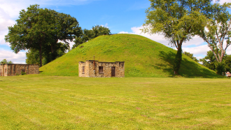 Picture of the Grave Creek Mound in Moundsville Ohio with a stone building in front of it.